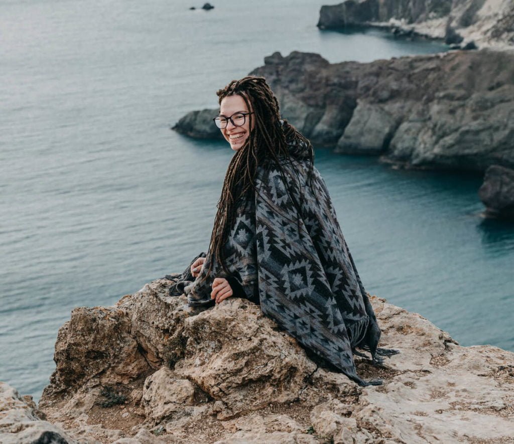 a person with long hair sitting on a rock by the water